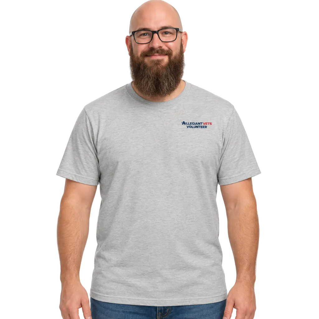 A man with a bald head, glasses, and a full beard wearing a light gray heather crewneck t-shirt with a small 'AllegiantVETS Volunteer' logo on the left chest, paired with blue jeans, against a plain white background.