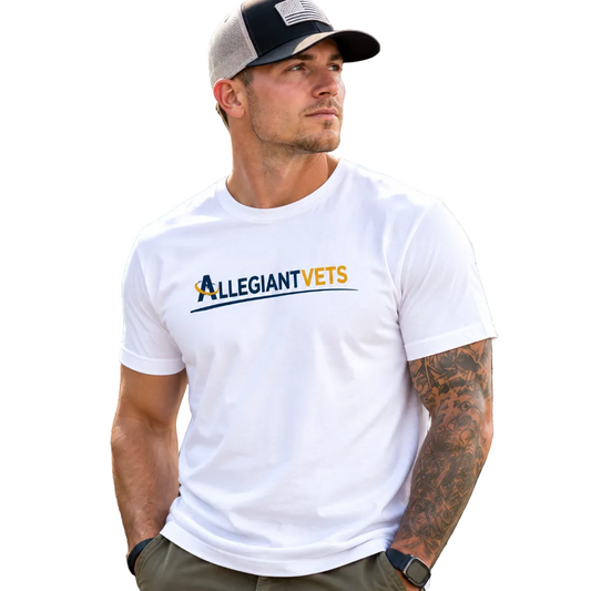 A man wearing a white crewneck t-shirt with the 'ALLEGIANTVETS' logo printed across the chest, paired with a black and gray trucker hat and khaki pants, standing outdoors against a dark fence.