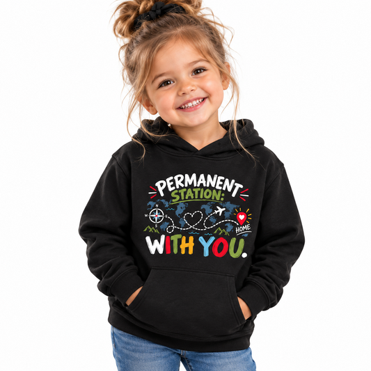A young girl with her hair in a high bun smiles while wearing a black hoodie with a colorful graphic that reads 'PERMANENT STATION: WITH YOU.' on the front, paired with blue jeans, against a plain white background.