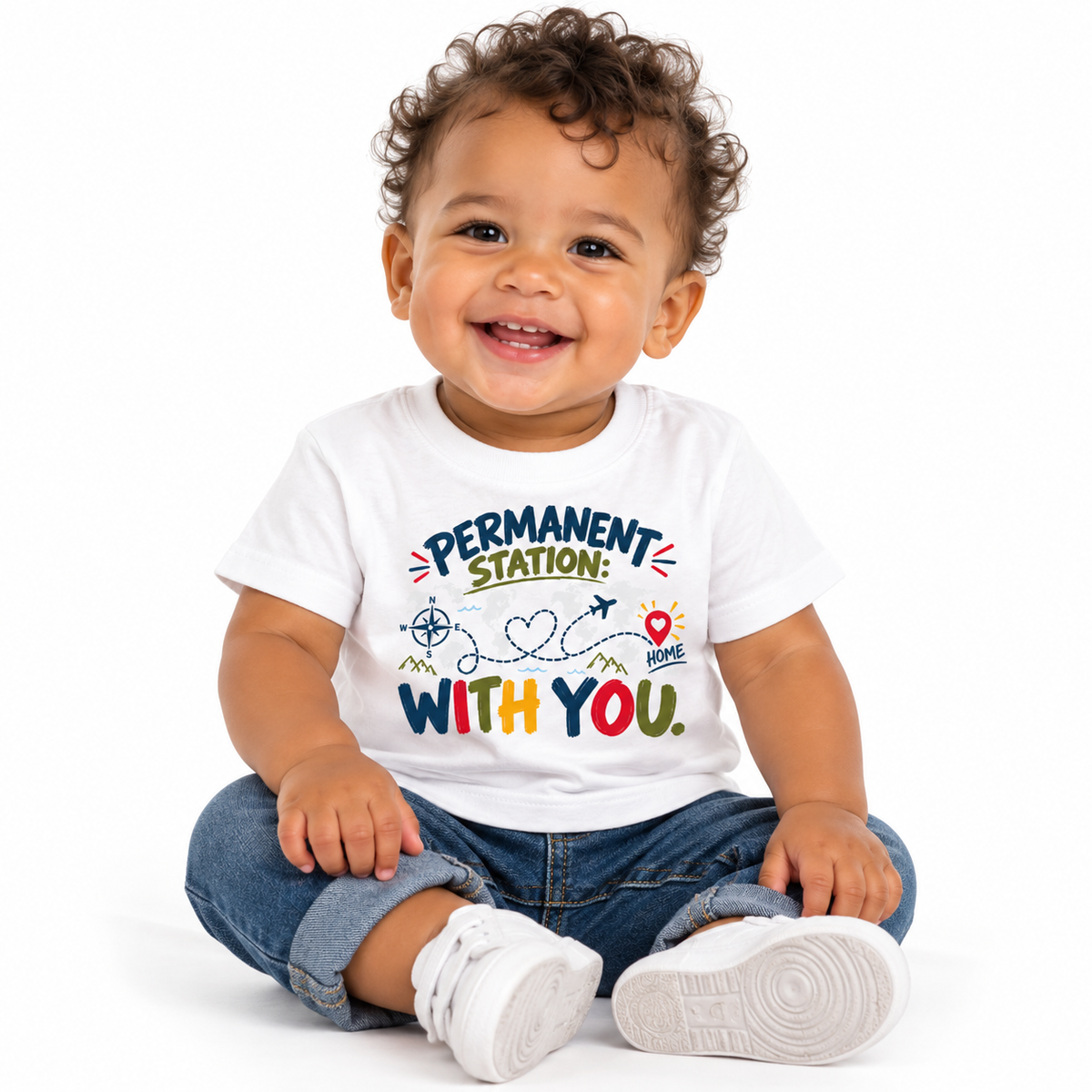 A smiling baby sitting cross-legged on a white studio background, wearing a white graphic T-shirt with the text 'PERMANENT STATION: WITH YOU.' and colorful travel-themed illustrations, along with blue jeans and white sneakers.